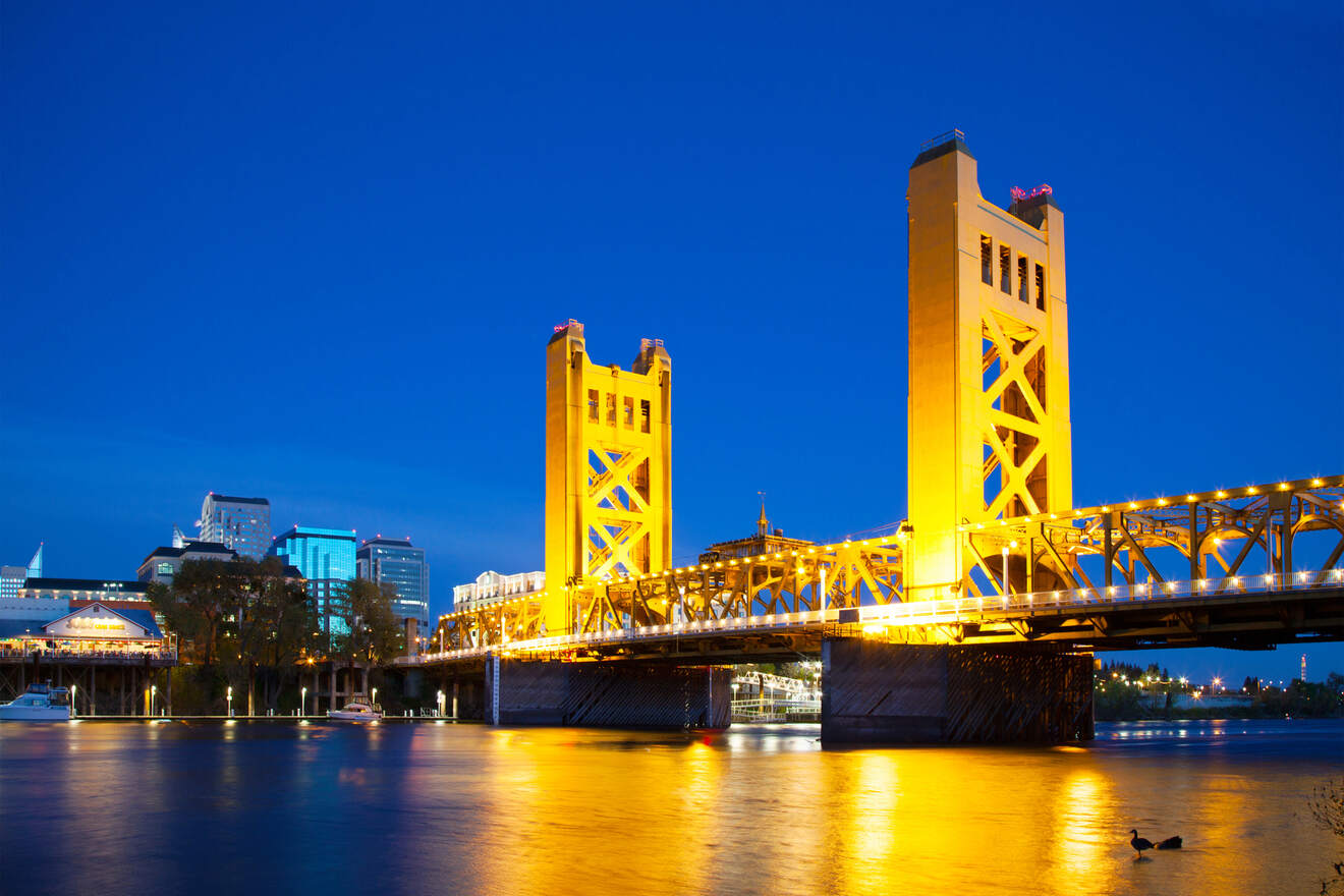 The iconic Tower Bridge in Sacramento illuminated at dusk, with its golden lights reflecting on the calm river water, creating a picturesque evening scene.